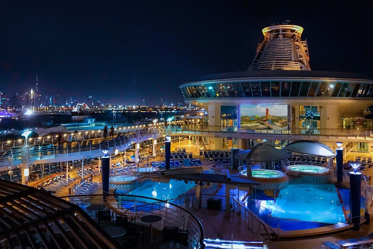 Cruise ship pool deck at night with illuminated pools, lounge seating, and ocean views